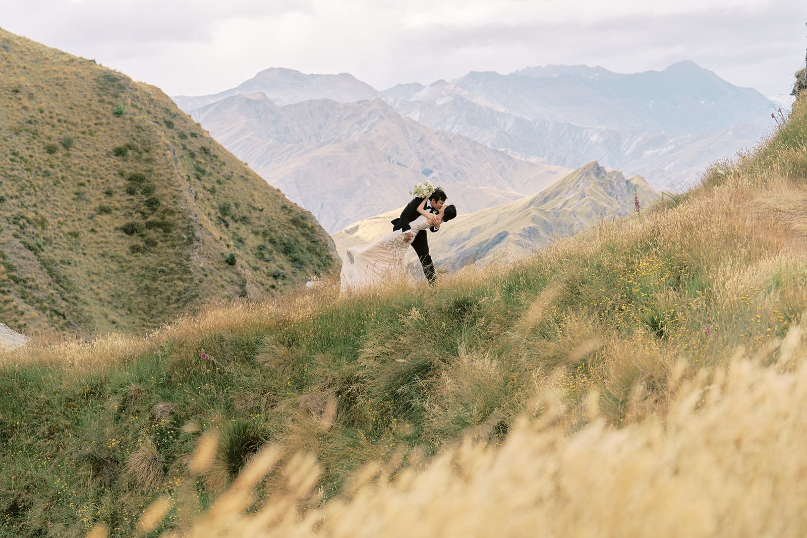 Queenstown New Zealand Heli Wedding Elopement Photographer クイーンズタウン　ニュージーランド　エロープメント 結婚式 | A groom carrying a bride through a grassy hillside with mountains in the background, captured by YURI, a Queenstown Wedding Photographer.